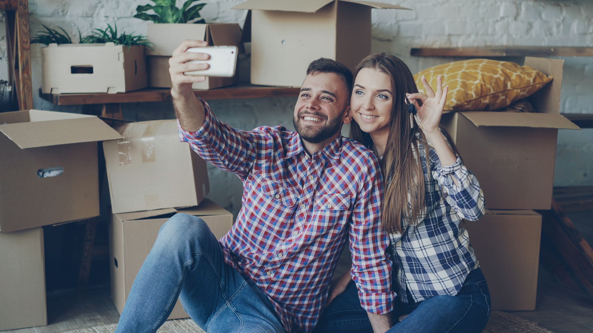 Paar sitzt lächelnd zwischen Umzugskartons und macht ein Selfie, in der Hand die neuen Wohnungsschlüssel